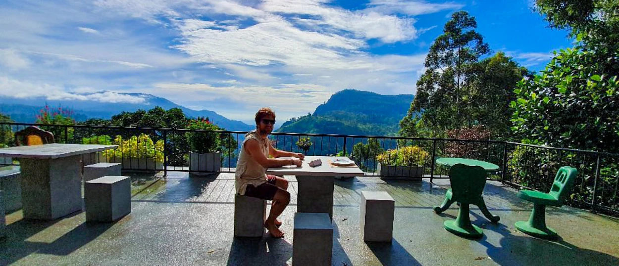 A person relaxes at a table on a terrace, enjoying a panoramic view of Udawalawe's green mountains.