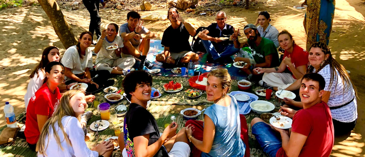 A happy group of travelers enjoys a communal outdoor lunch under the shade of trees in Zanzibar.