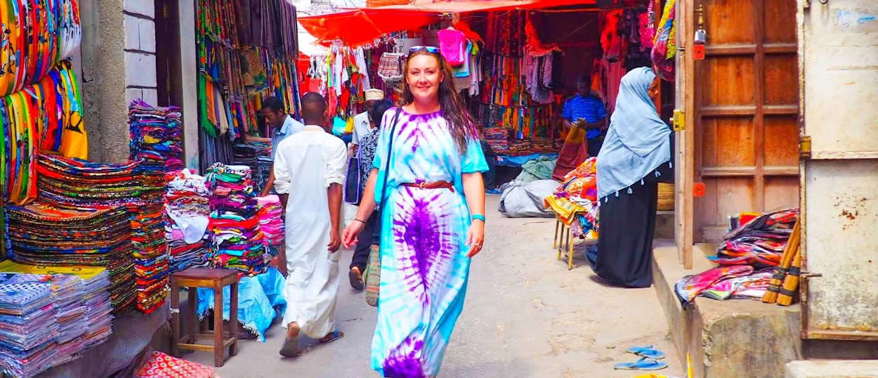 A smiling woman in a colorful tie-dye dress walks through a vibrant market in Zanzibar.