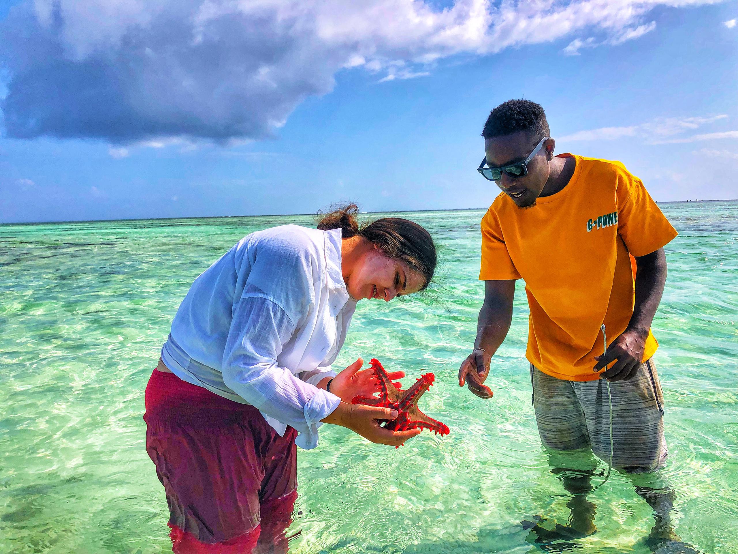 A volunteer and a member of the local team hold a red starfish in the shallows.