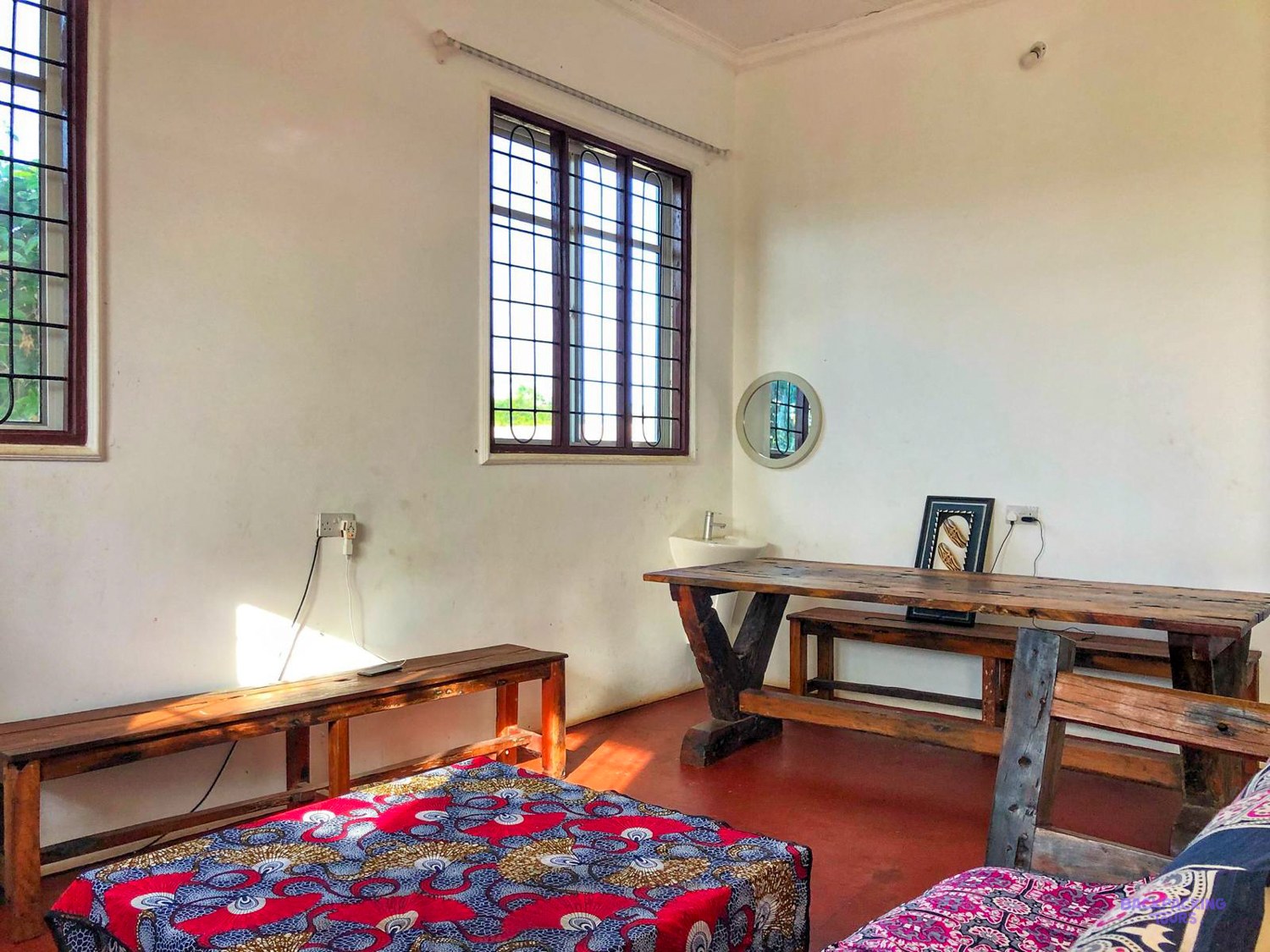 A long wooden desk and bench stand under the barred window of a standard room.