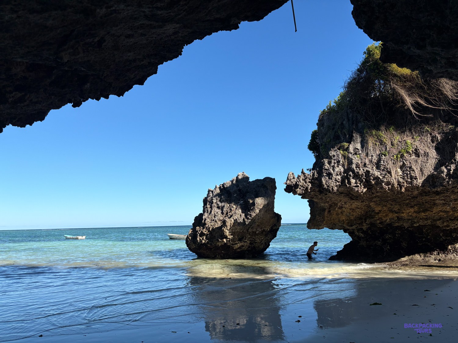 A swimmer stands in the shallows beneath an undercut coral cliff and a sea stack.