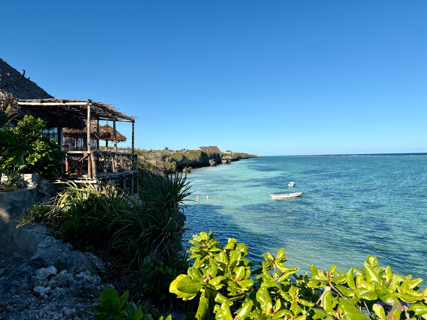 A thatched wooden bar stands on the low cliff above the turquoise reef and a moored boat.