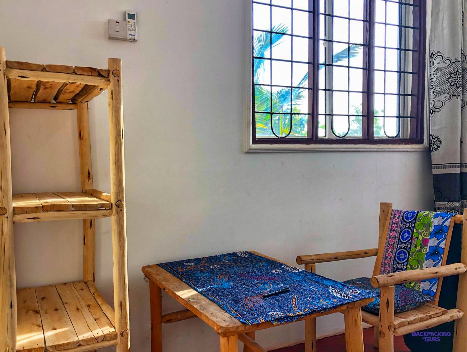 A wooden table and chair sit by a window looking out onto palm leaves.