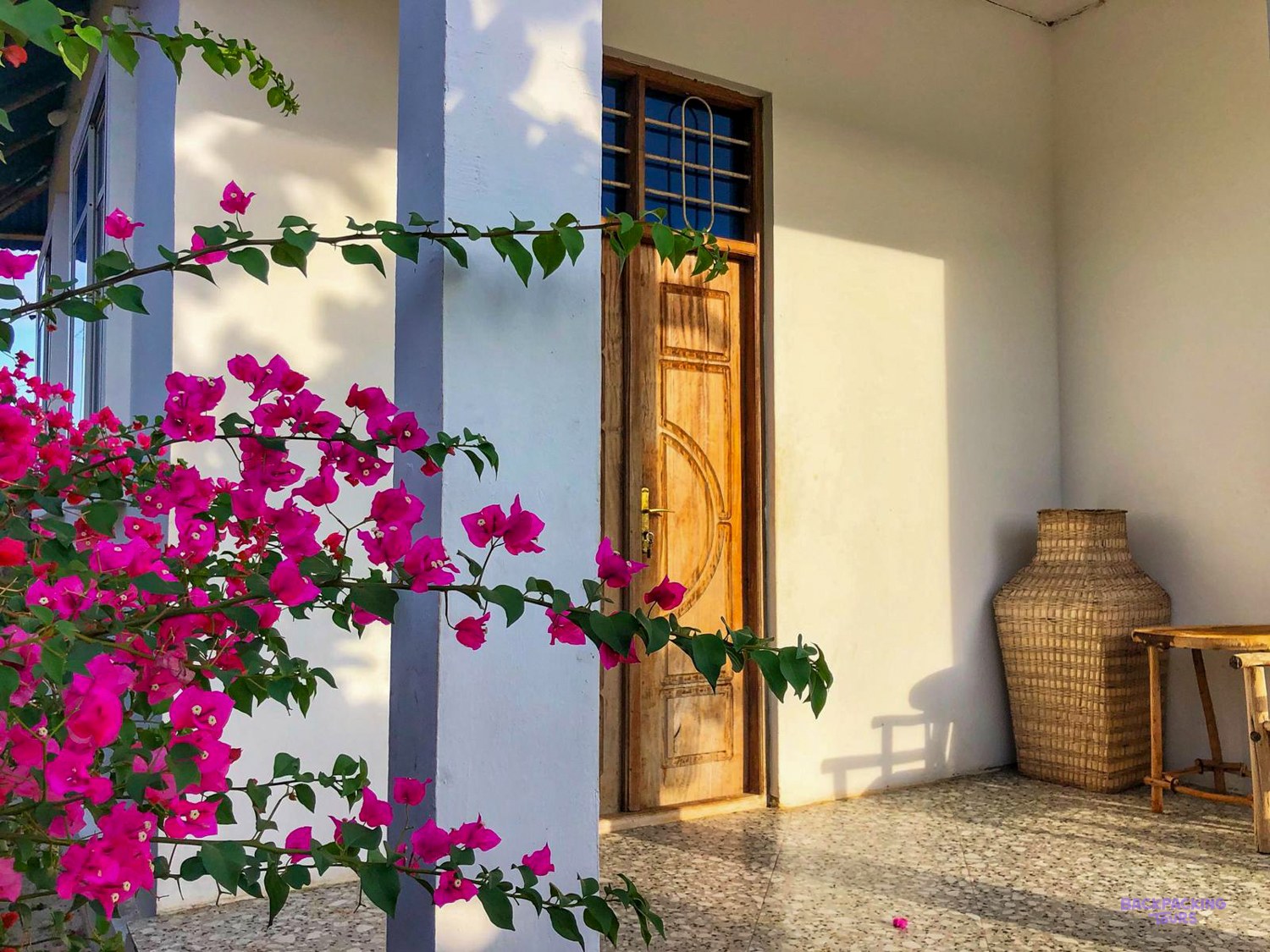 Pink bougainvillea frames a carved wooden door on the shaded veranda of the house.
