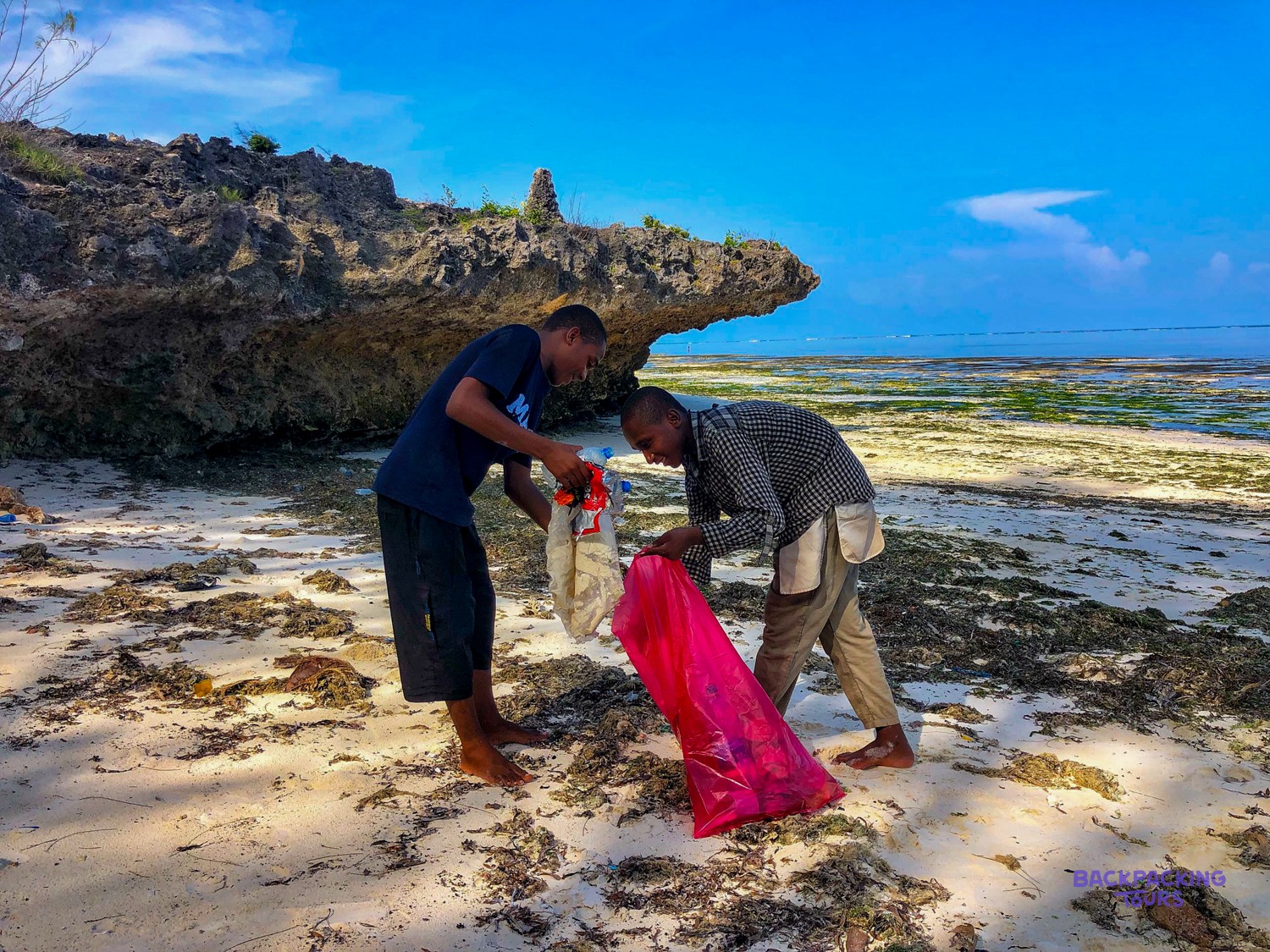 Two men hold open a pink sack and clear litter from the rocky shoreline.