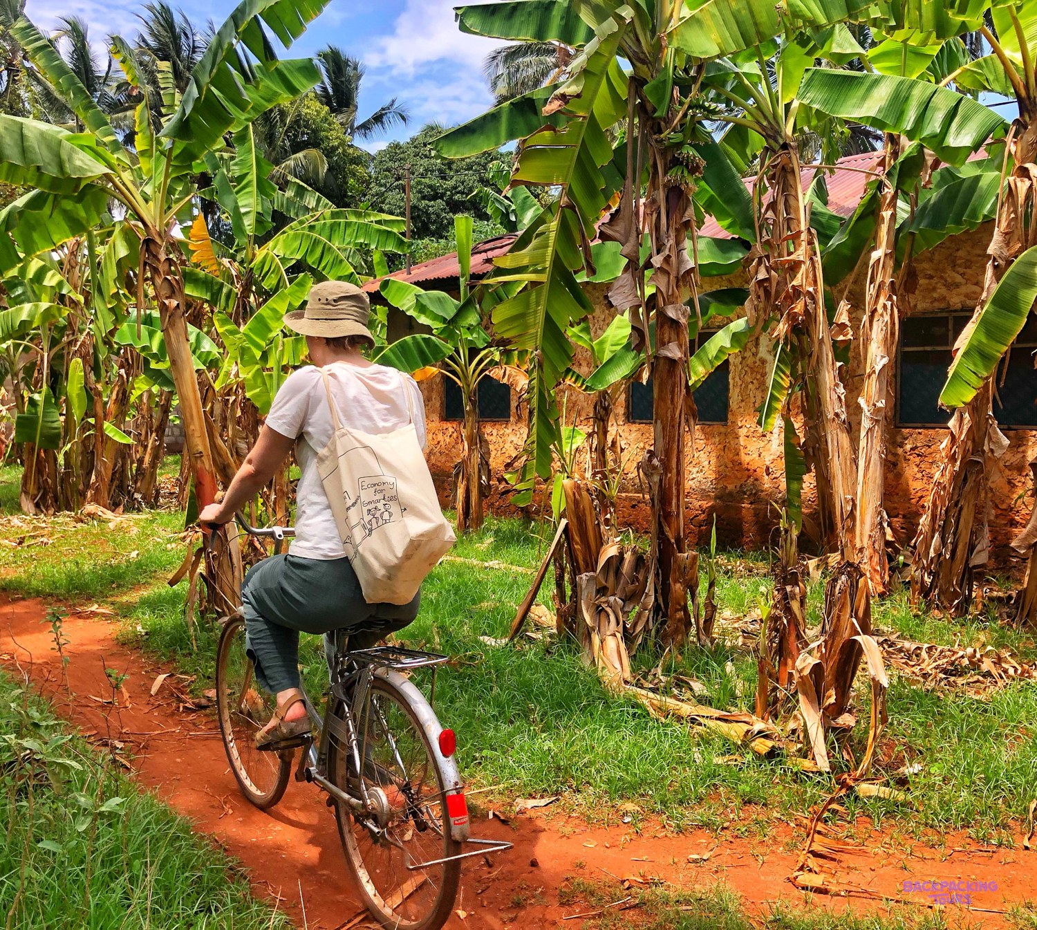 A volunteer cycles a red earth path through banana trees past a village house.