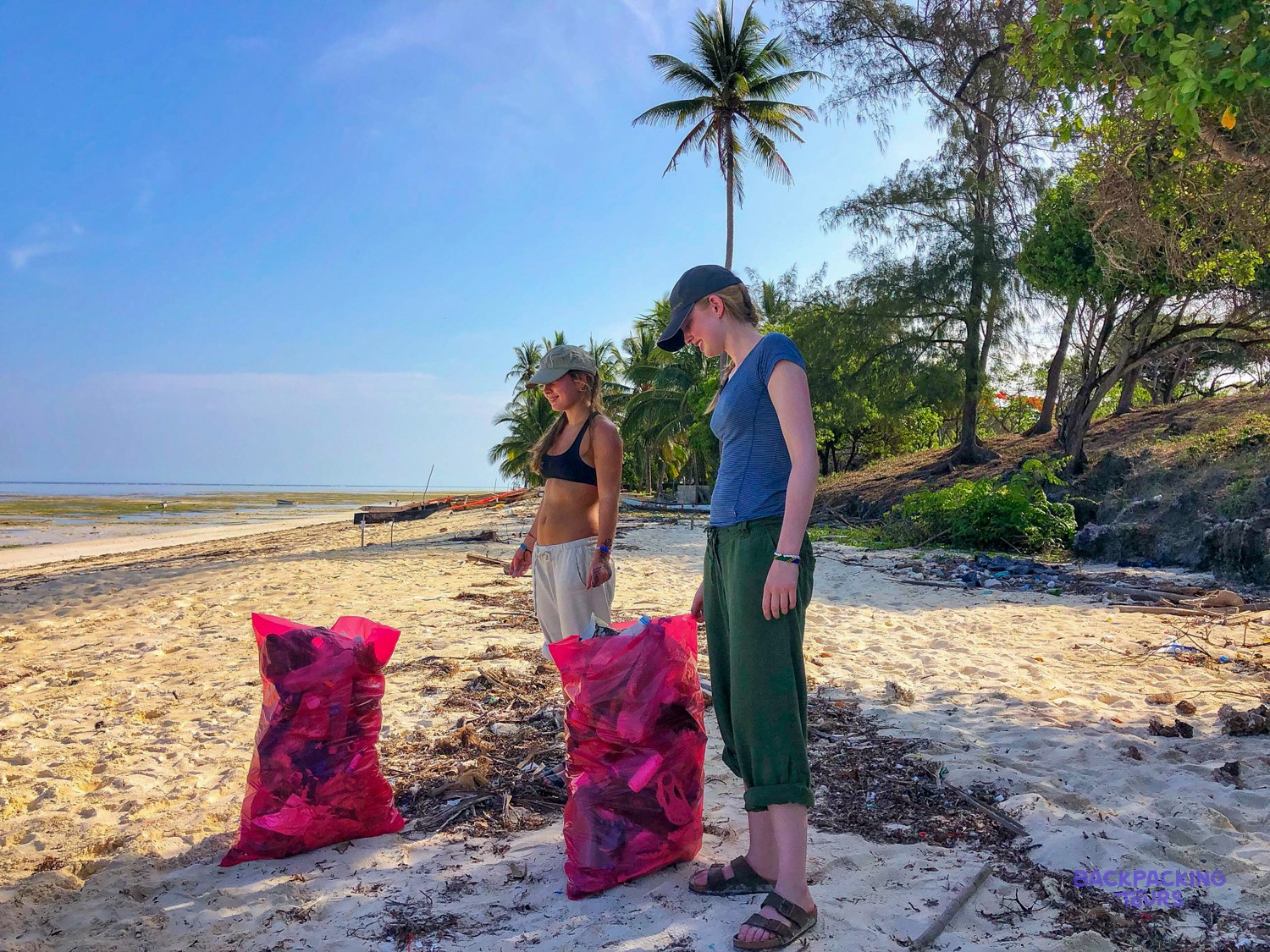 Two volunteers stand over full pink rubbish sacks on the sand after a beach clean.