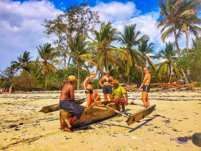 A group gathers around a wooden outrigger canoe on the sand beneath tall coconut palms.