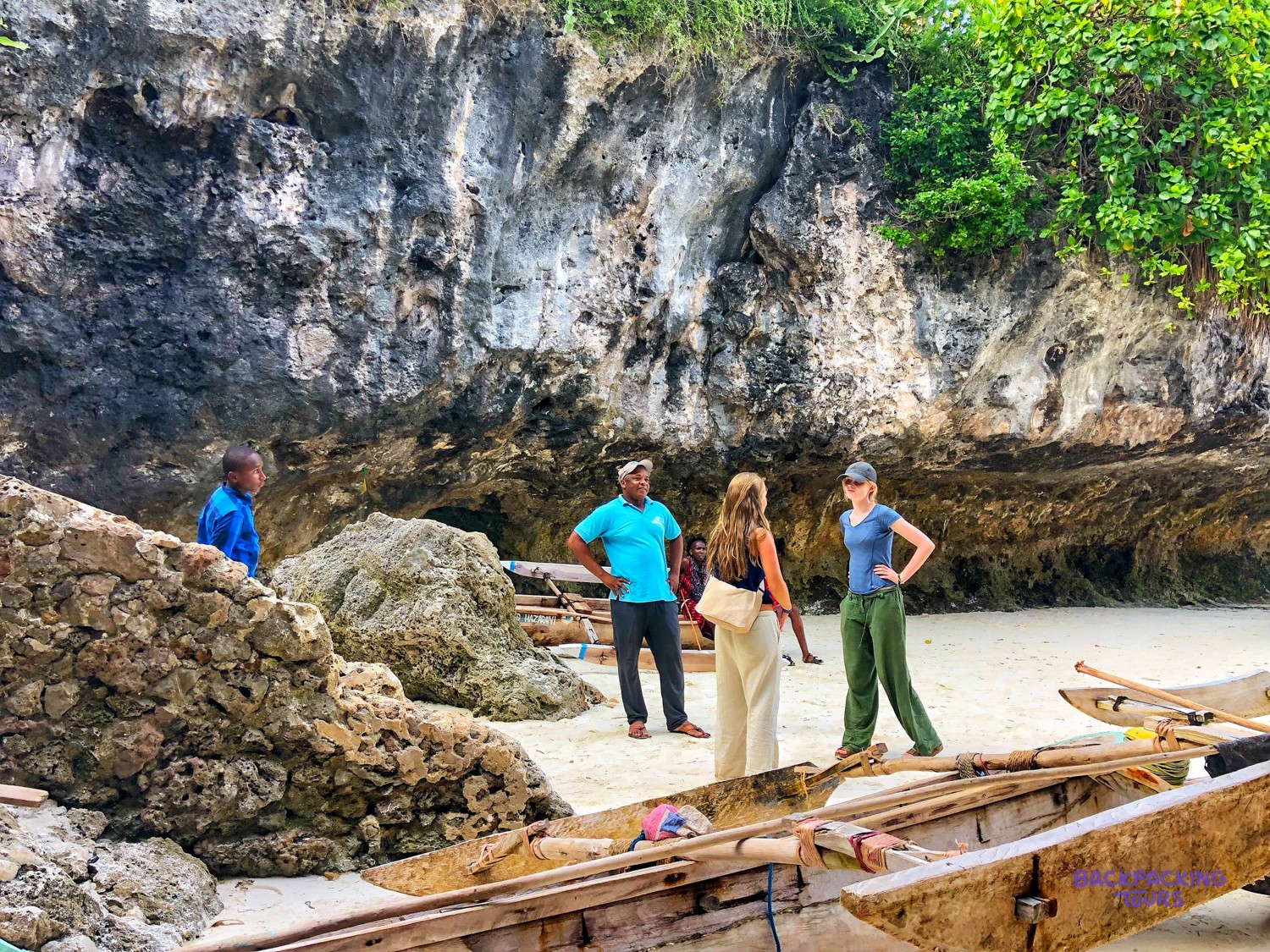 A guide and two volunteers stand among outrigger canoes on the sand below a coral cliff.