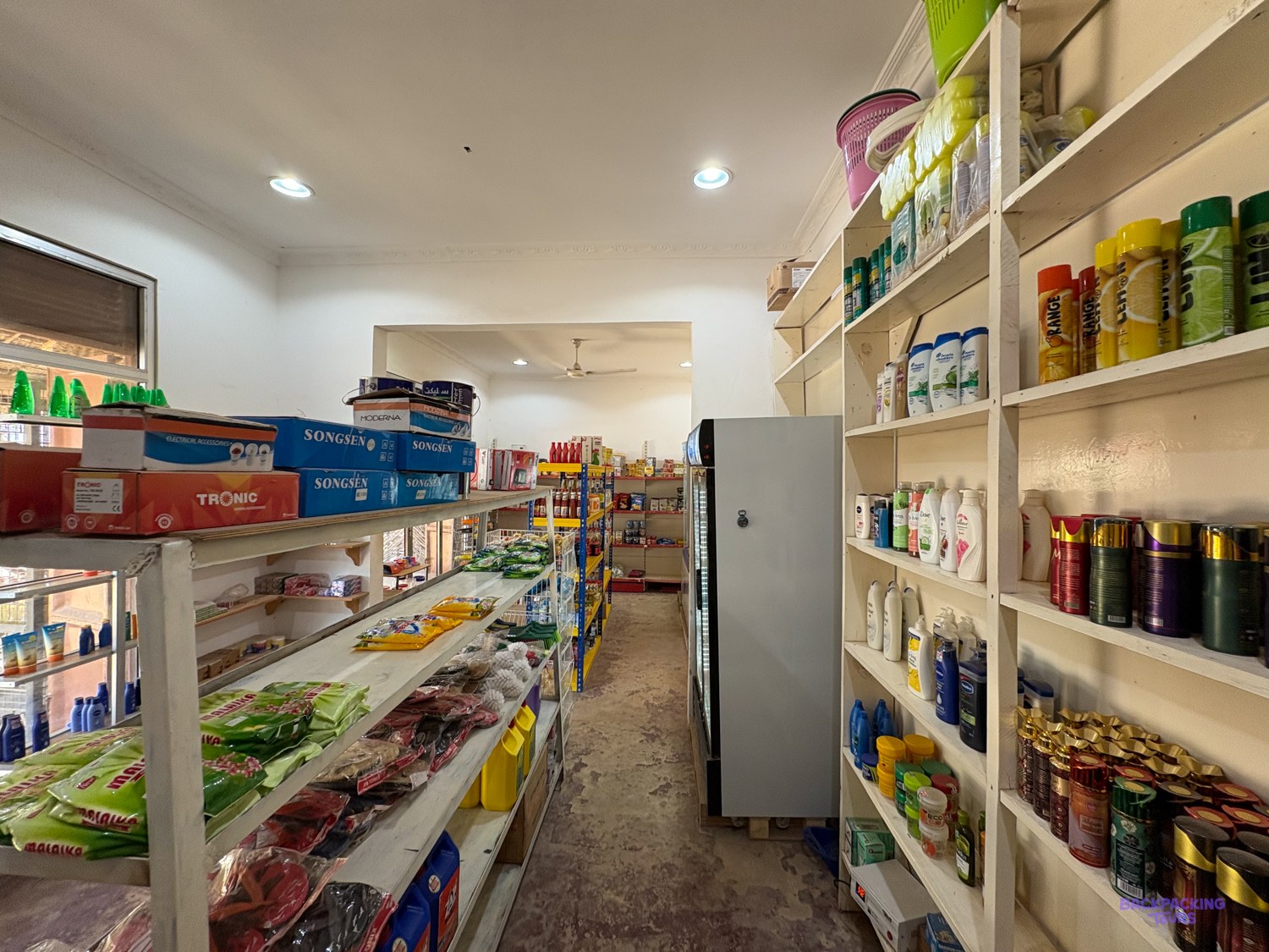 An aisle of household goods and toiletries runs the length of a village shop.