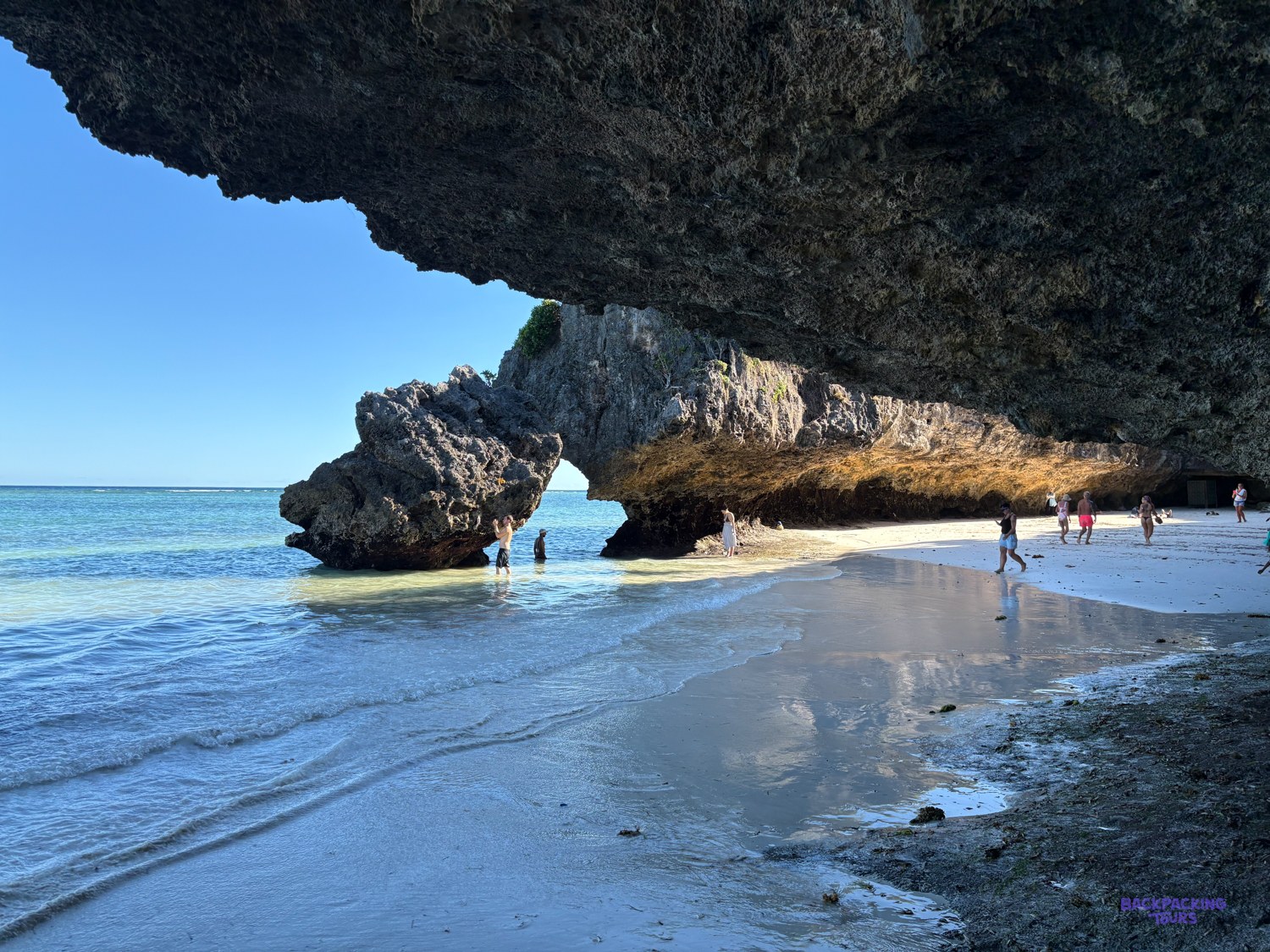 People wade and walk the wet sand beneath a wide coral arch at low tide.