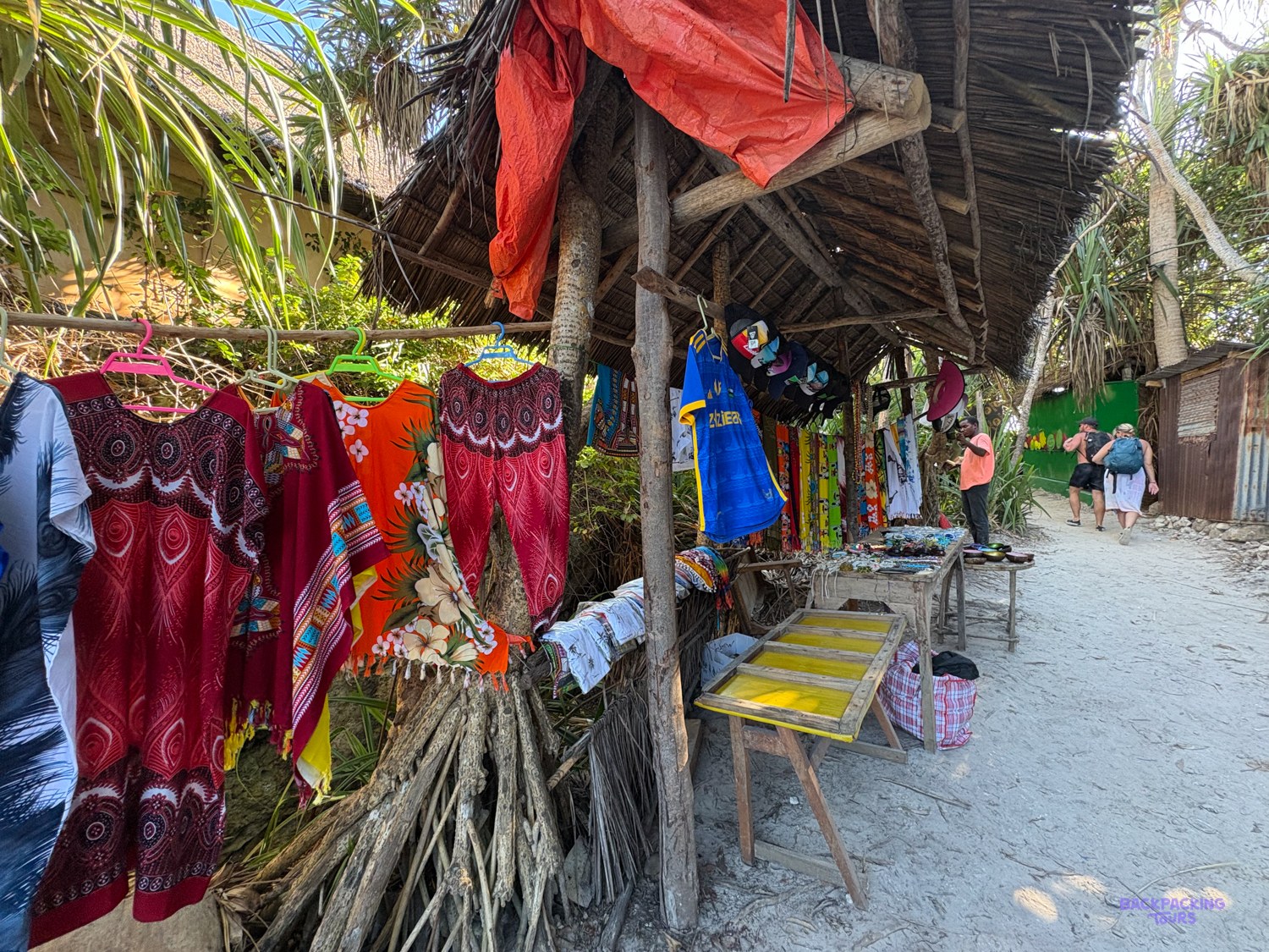 Dresses and printed cloth hang from a thatched craft stall on a sandy beach path.