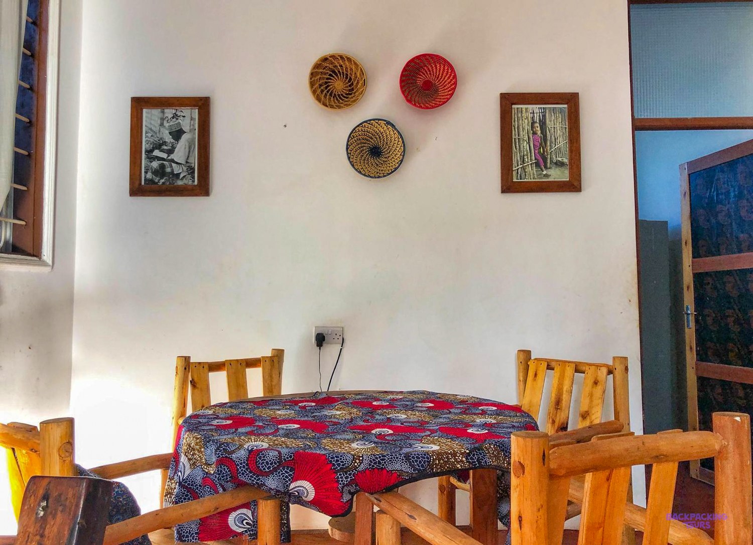 Woven baskets and framed photographs hang above a round table laid with printed cloth.