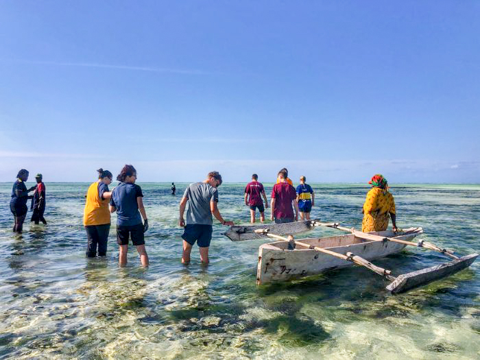 The whole group stands in the shallows around an outrigger canoe at low tide.