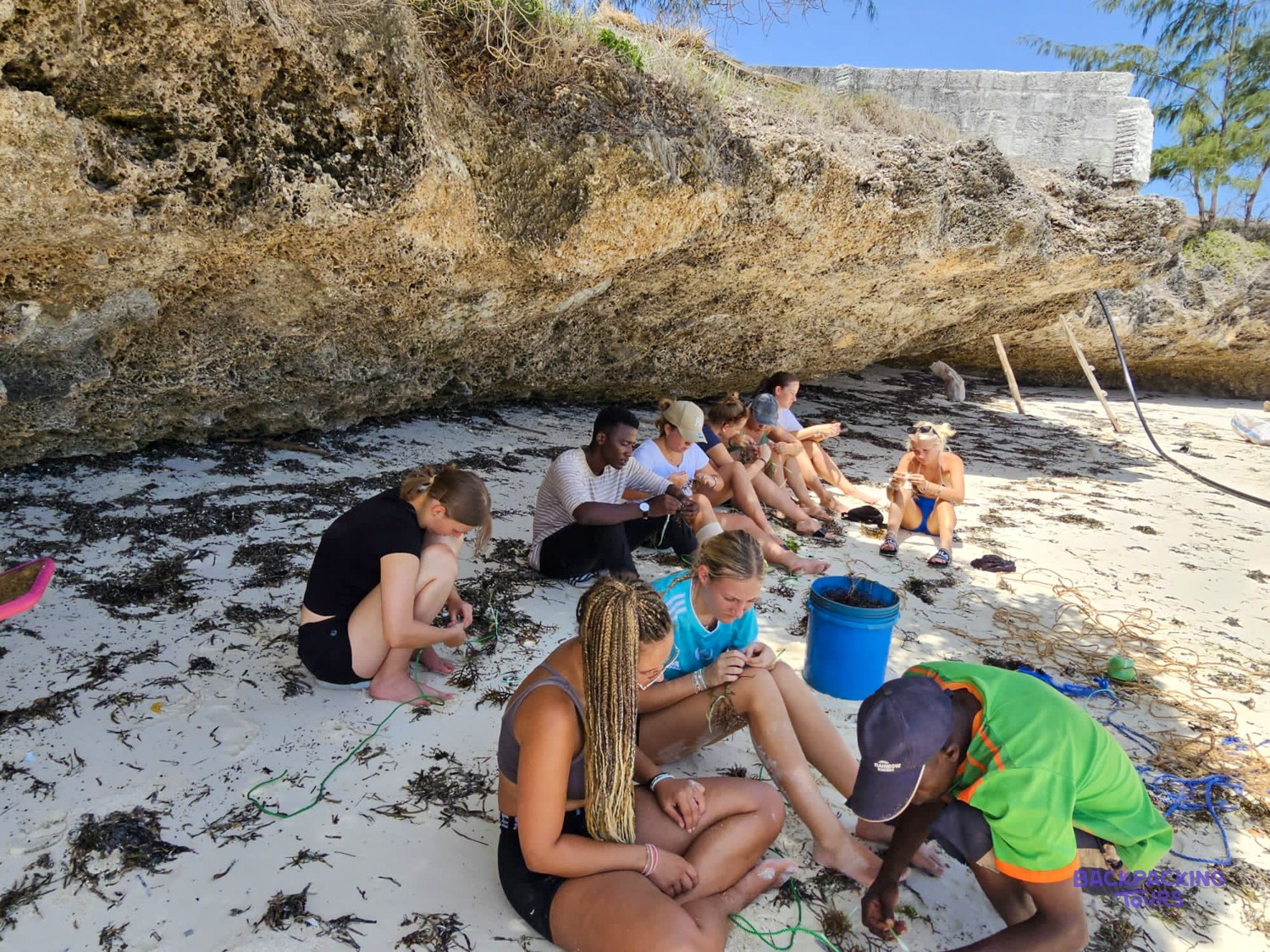 The whole group sits in the shade of the coral cliff, working with seaweed and rope.