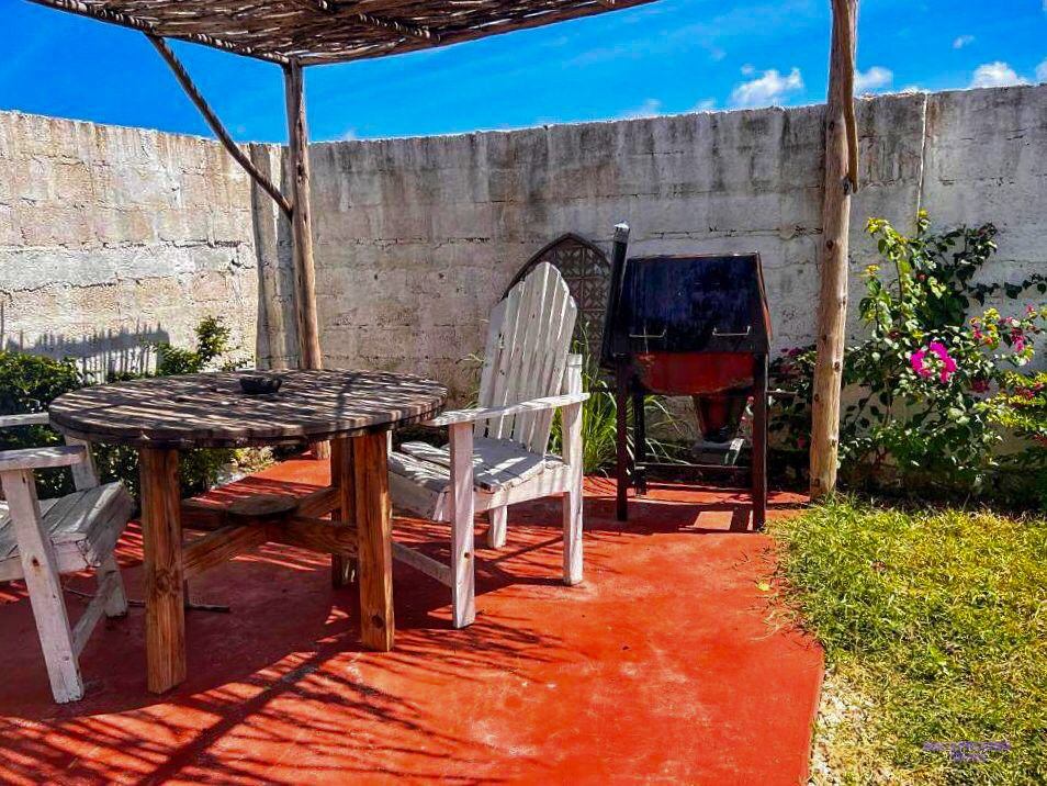 A round table and chairs sit under a wooden pergola beside a barbecue drum.