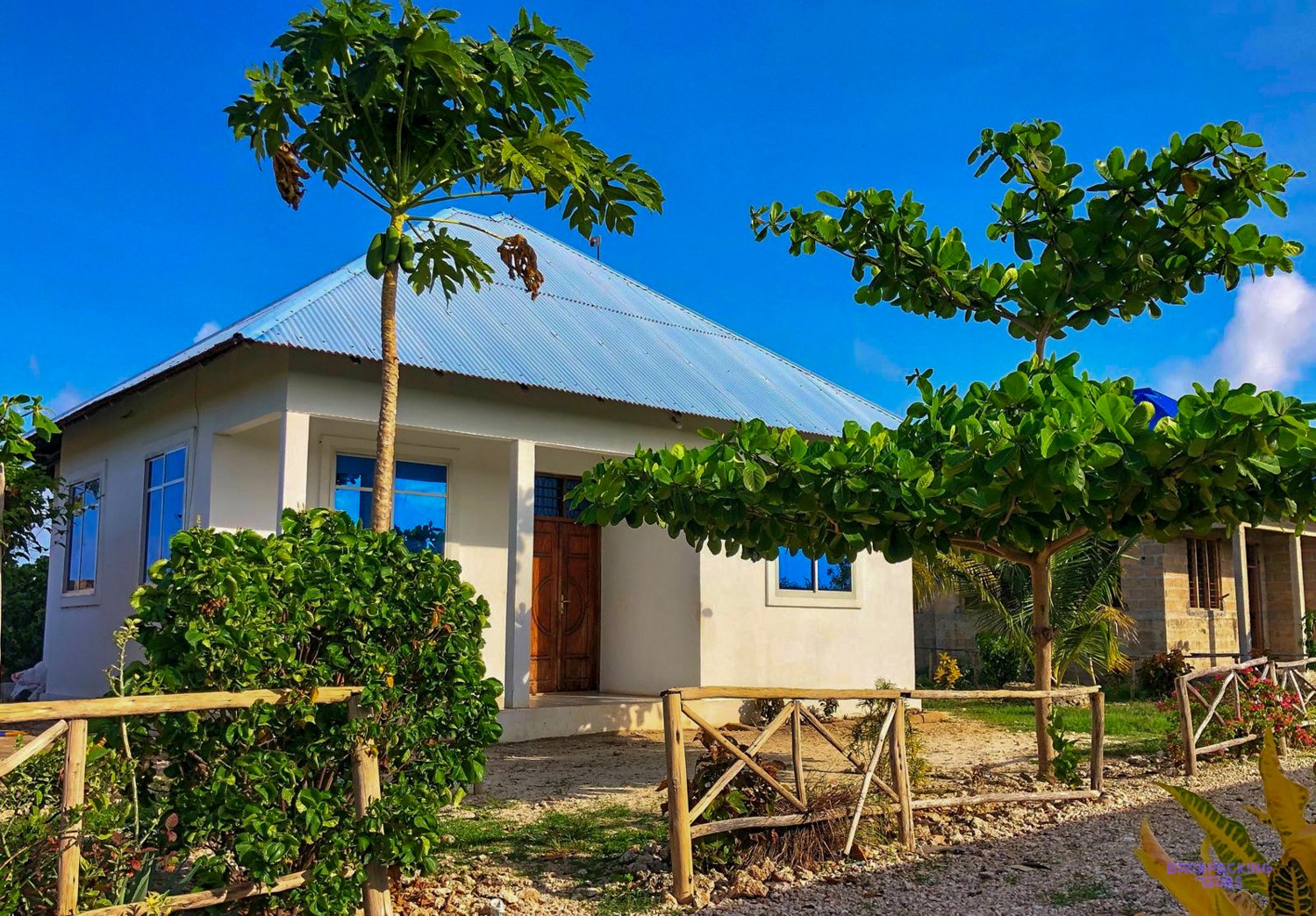 A white volunteer house with a blue tin roof stands behind a rustic garden fence.