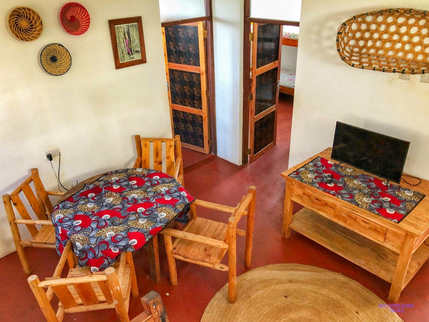 Wooden chairs, a printed cloth on the table and a television furnish the living room.