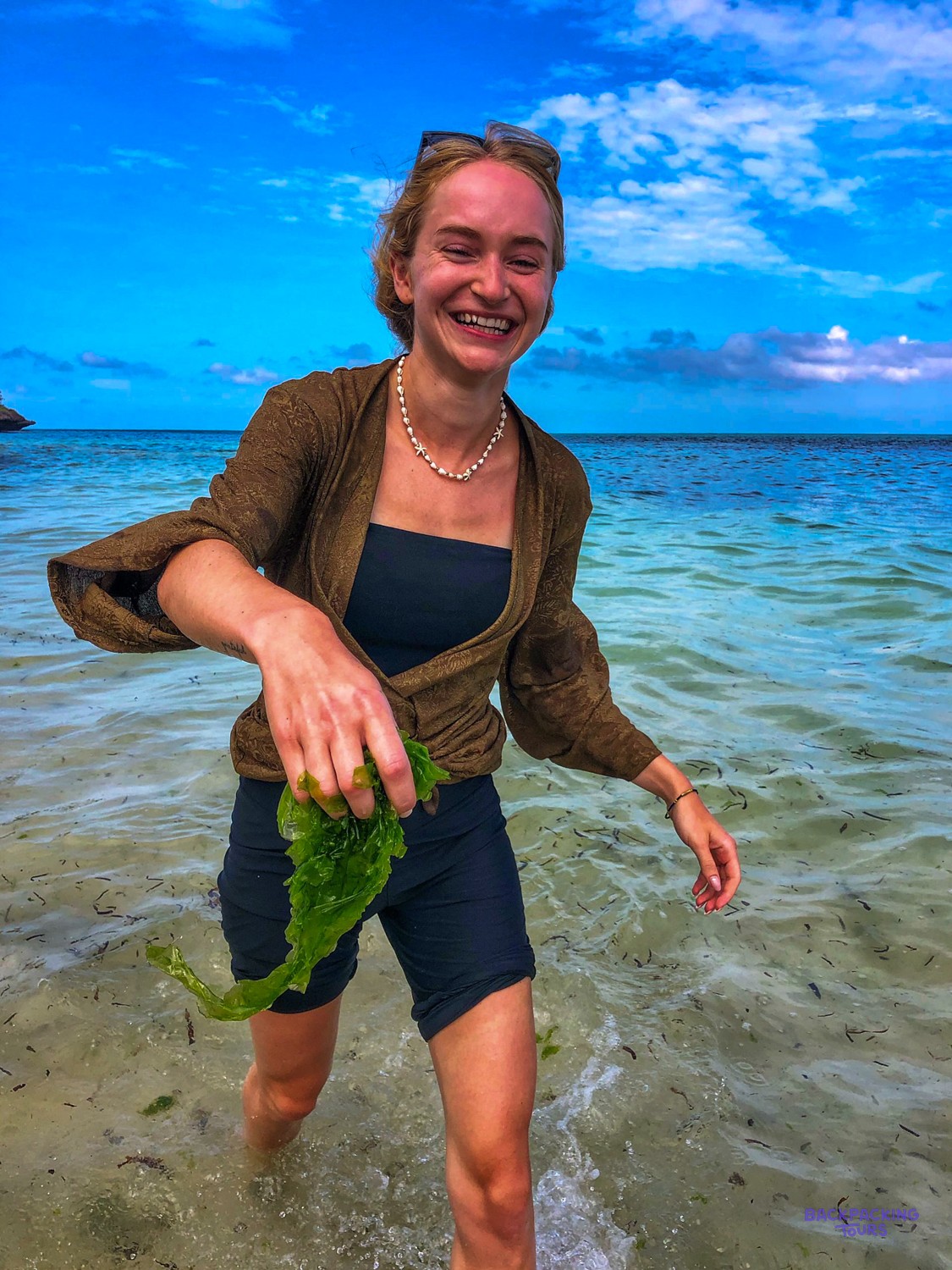 A smiling volunteer wades out of the sea holding a handful of freshly cut seaweed.
