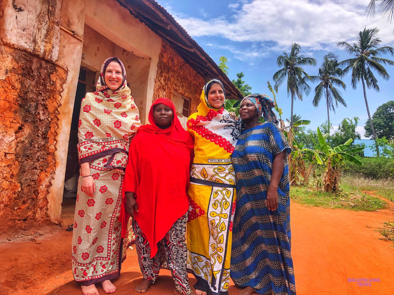 Two volunteers wrapped in patterned kanga stand with two village women outside a house.
