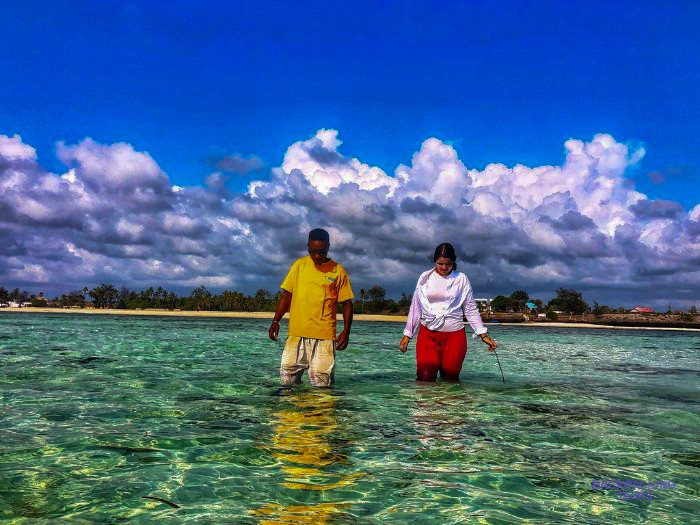 Two of the local team wade through clear turquoise shallows under a tall cloudy sky.