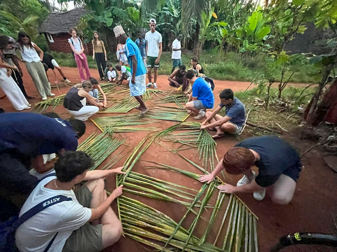 Volunteers and villagers sit on the red earth weaving long palm fronds into matting.