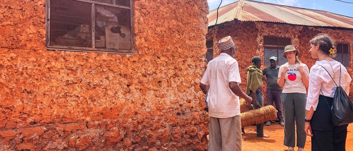 A guide and two volunteers stand beside the coral rag wall of a village house.