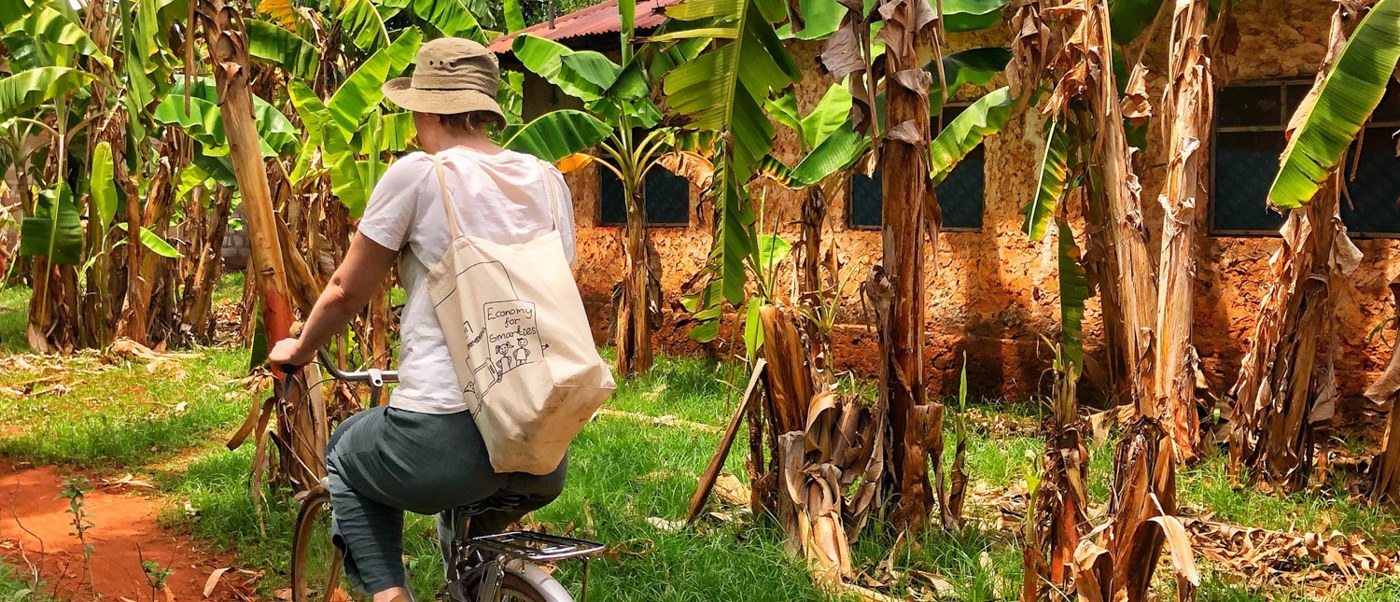 A volunteer cycles a red earth path through banana trees past a village house.