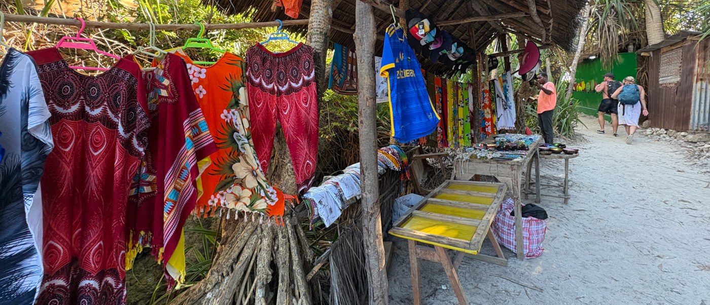Dresses and printed cloth hang from a thatched craft stall on a sandy beach path.