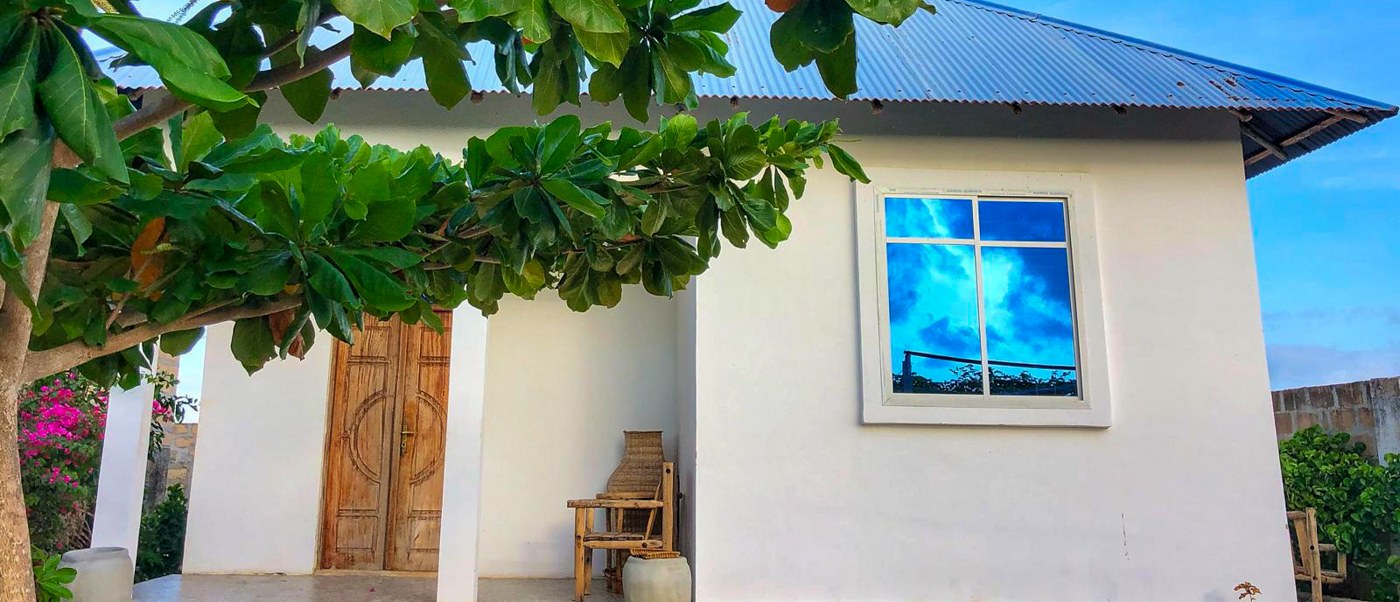 A white house with a blue roof and a carved door sits under a spreading almond tree.