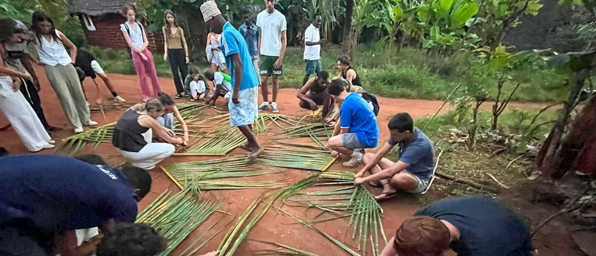 Volunteers and villagers sit on the red earth weaving long palm fronds into matting.
