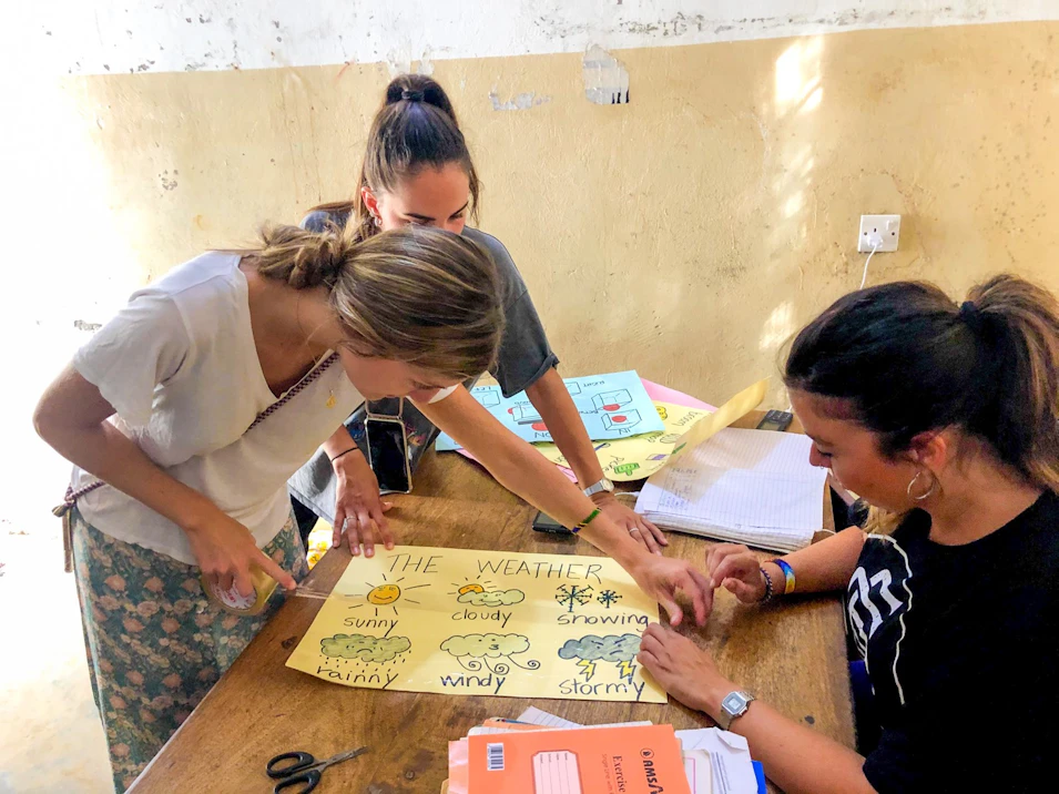 Three women volunteers collaboratively teaching about weather using a handmade poster in Zanzibar.