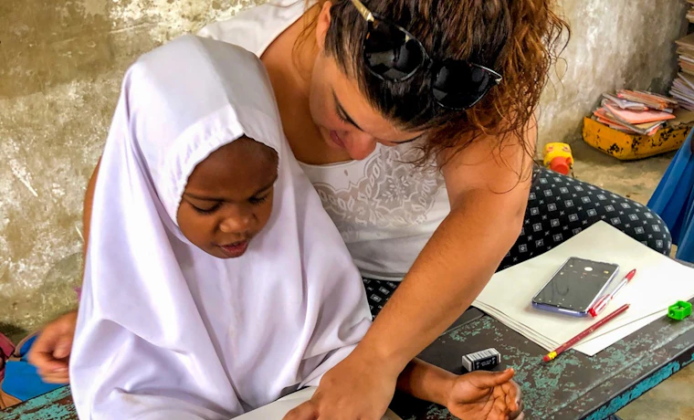 A volunteer teacher helps a young child practice writing numbers at a worn desk in Tanzania.