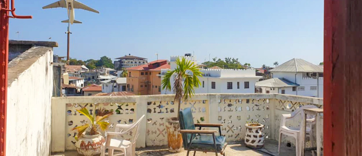 Rooftop terrace in Zanzibar with chairs, potted plants, and a model airplane above city buildings.