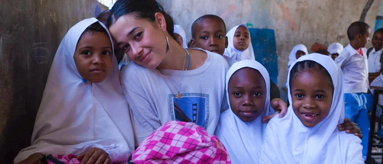 A smiling female volunteer poses closely with young students wearing white head coverings in a Zanzibar classroom.