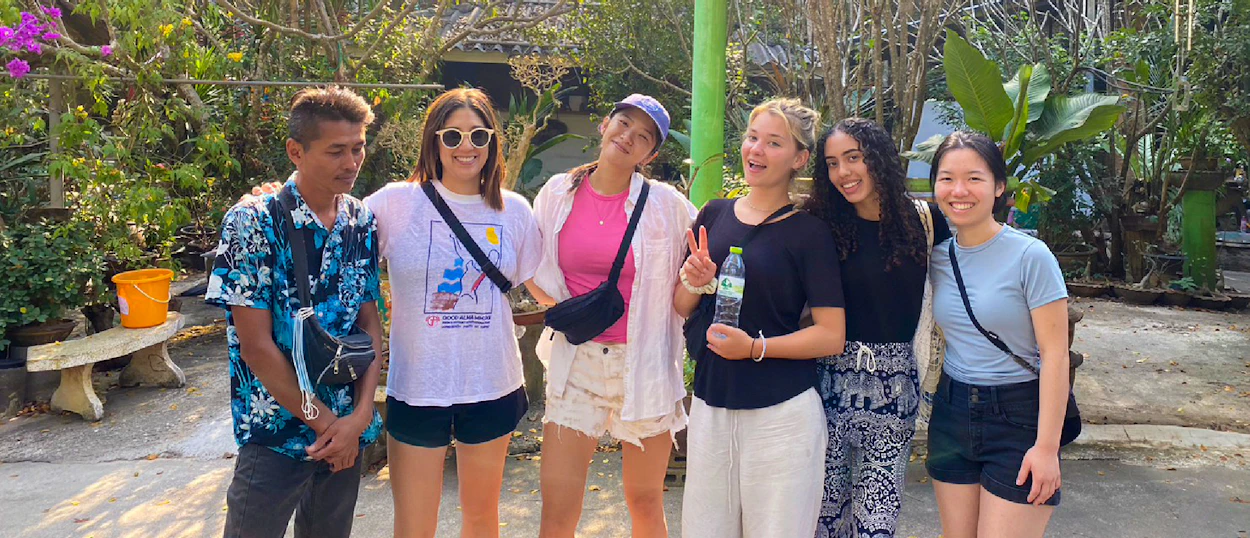 Six happy volunteers and a local guide pose together in a lush outdoor setting in Thailand.