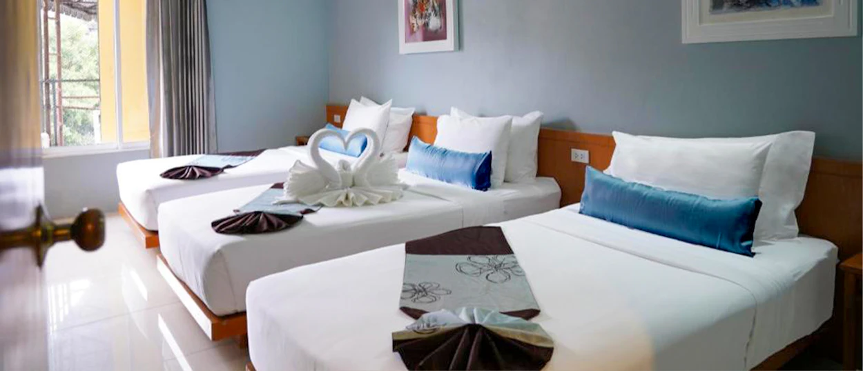 A bright hotel room in Bangkok with three single beds, white linens, and blue pillows.