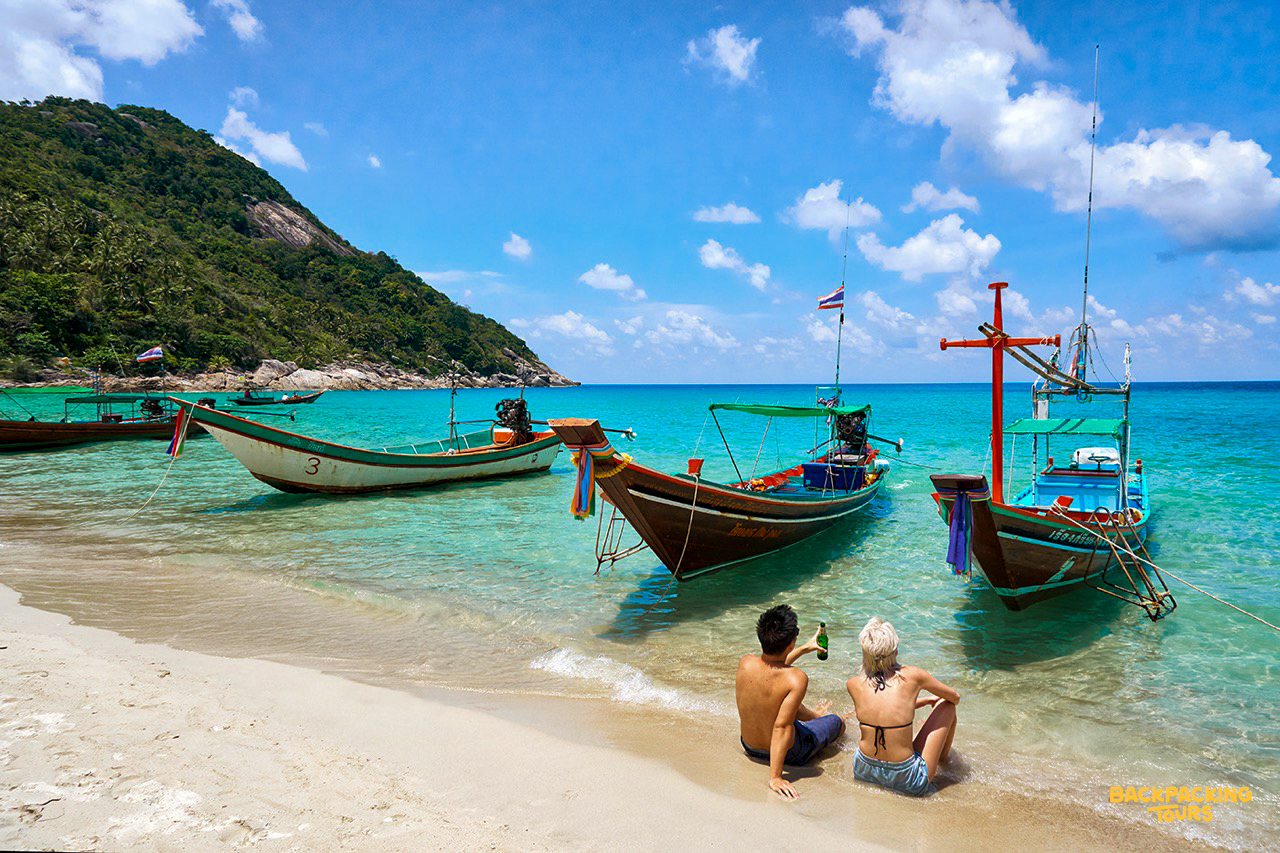Longtail boats on a sandy beach with turquoise waters on Koh Phangan where the backpacking tour group arrives at the tropical island