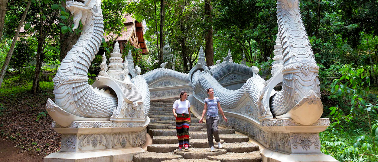 Two women ascend a temple staircase flanked by white naga statues amidst lush jungle in Chiang Mai.