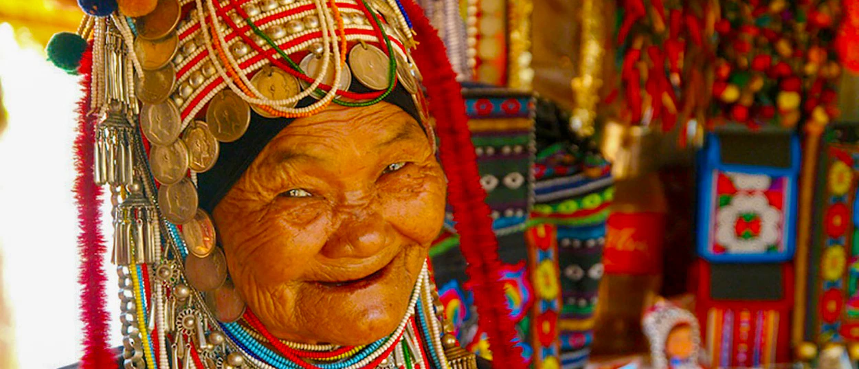 An elderly hill tribe woman in Chiang Mai smiles warmly, wearing traditional ornate headwear with coins and beads.