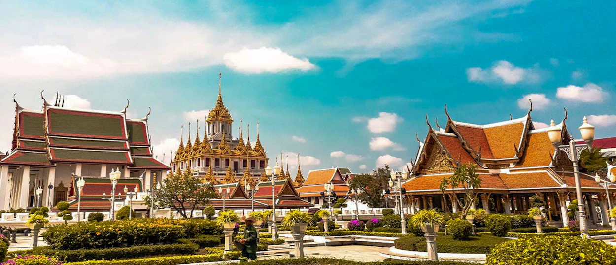 Ornate Thai temples with golden spires and tiered roofs in a beautiful garden under a bright blue sky.