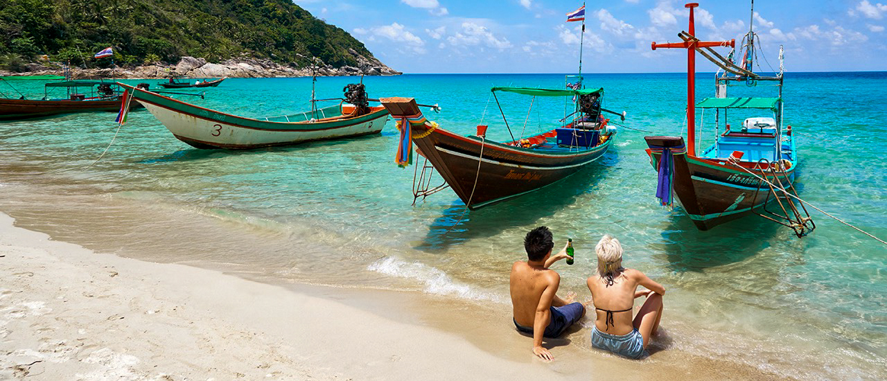 Backpacking tour friends relaxing on Koh Phangan beach with longtail boats anchored nearby