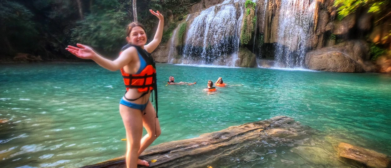 Joyful woman in a life vest on a log in emerald pools at Erawan Waterfalls, Thailand.