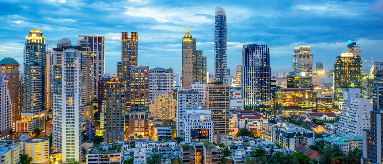 A vibrant Bangkok city skyline at dusk, featuring many illuminated skyscrapers against a deep blue sky.