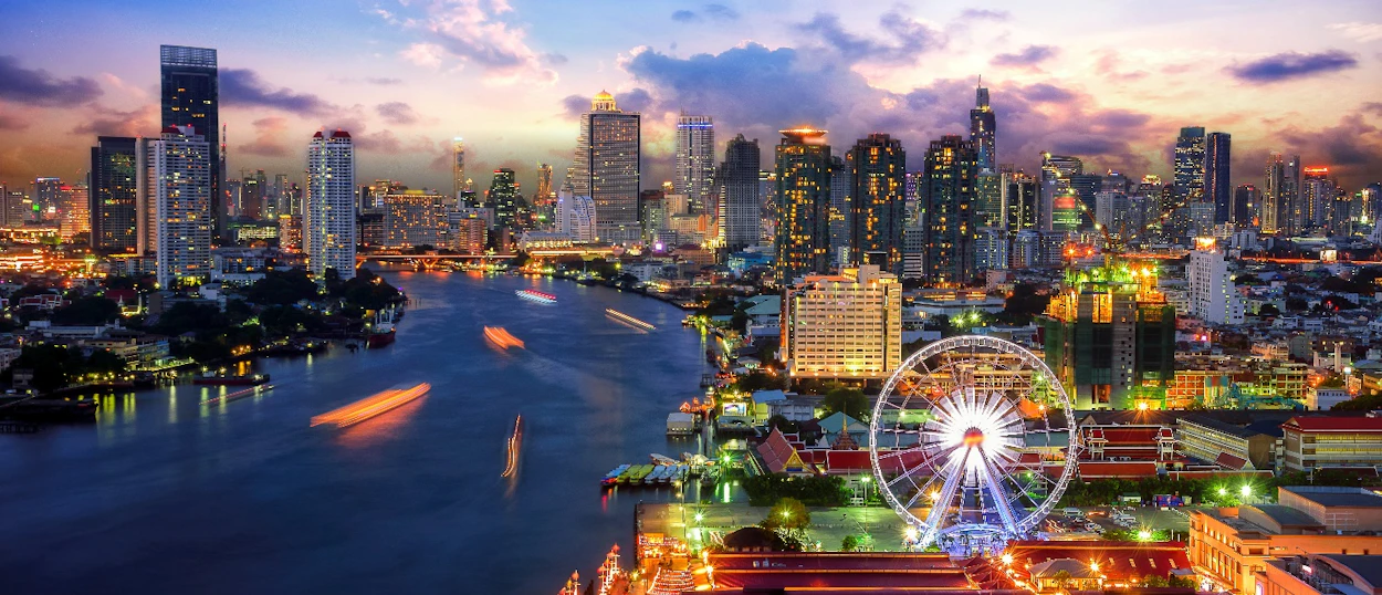 Panoramic view of Bangkok at twilight, featuring skyscrapers, a river with boat lights, and a Ferris wheel.