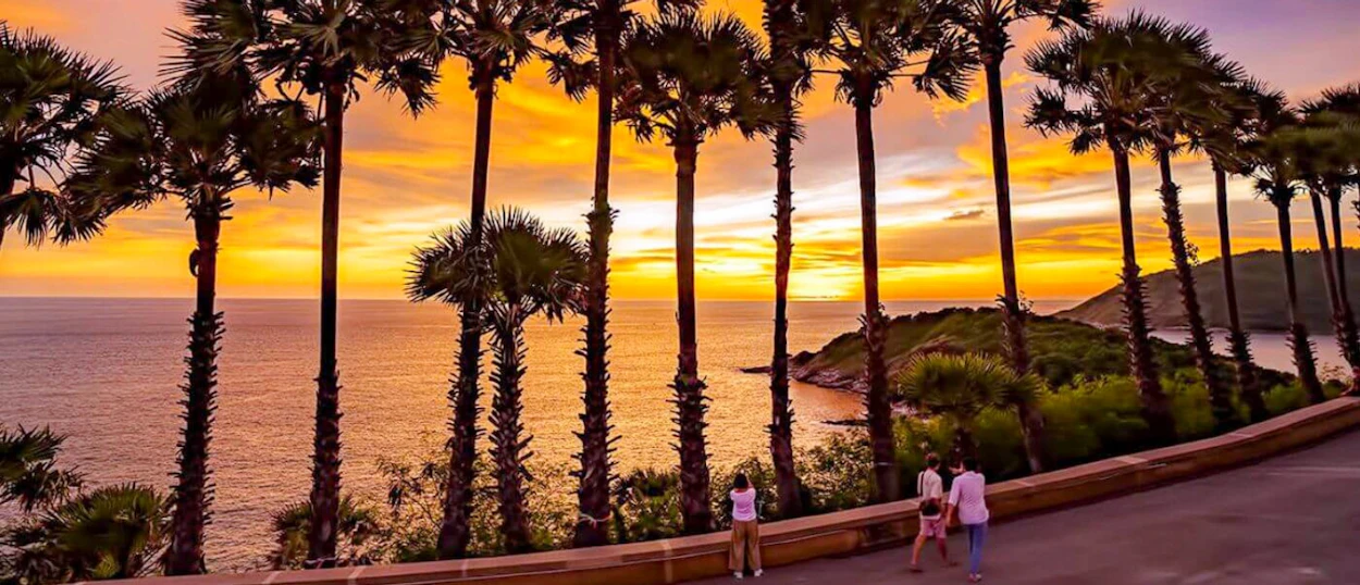 Tourists watch a vibrant sunset over the ocean, framed by tall palm trees in Phuket, Thailand.