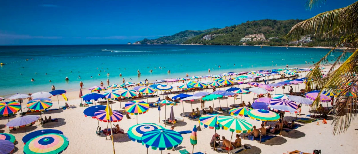 Colorful umbrellas line a beautiful tropical beach in Phuket with people swimming in turquoise water.