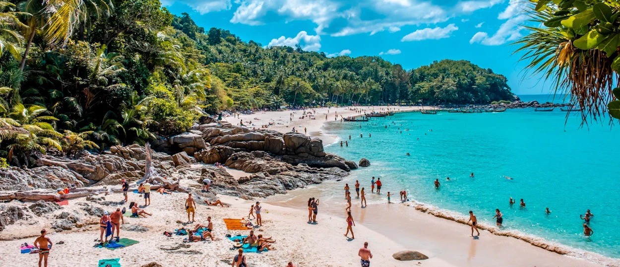 Crowded tropical beach in Phuket with many people swimming in turquoise water and relaxing on the sand.