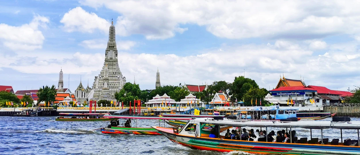 Colorful longtail boats navigate the Chao Phraya River with the majestic Wat Arun temple in Bangkok.